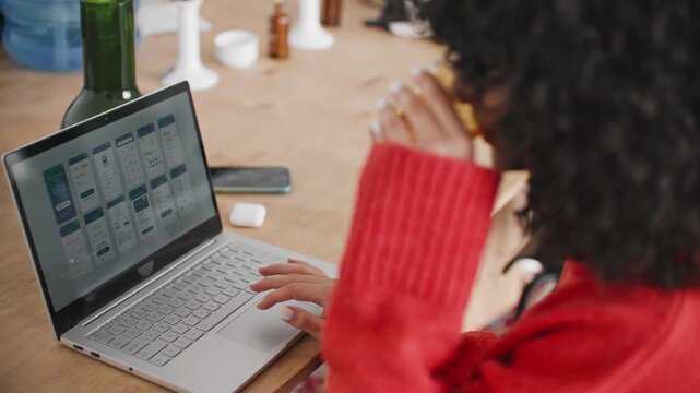 Over shoulder view of young woman using laptop while holding glass of tea at wooden table. Designer reviewing mobile app interface layouts. Working on digital product development in modern office.