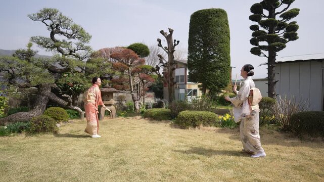 Two women in traditional robes walk through a manicured garden. Their movements are graceful, slow, and deliberate. They stroll past sculpted trees and a stone lantern. The scene evokes serenity, calm