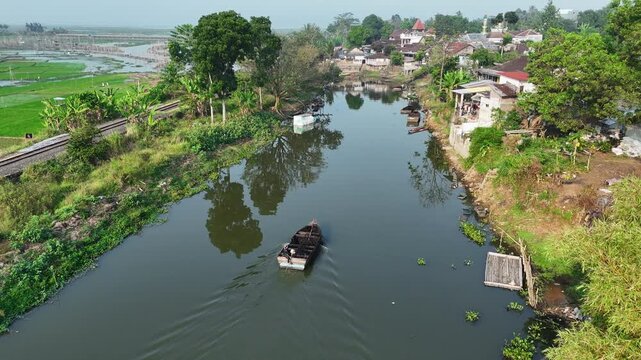 Aerial view of a small boat navigating the calm waters of Lake Rawa Pening, surrounded by lush greenery and railway tracks in Waduk Rawapening, Central Java, Indonesia.