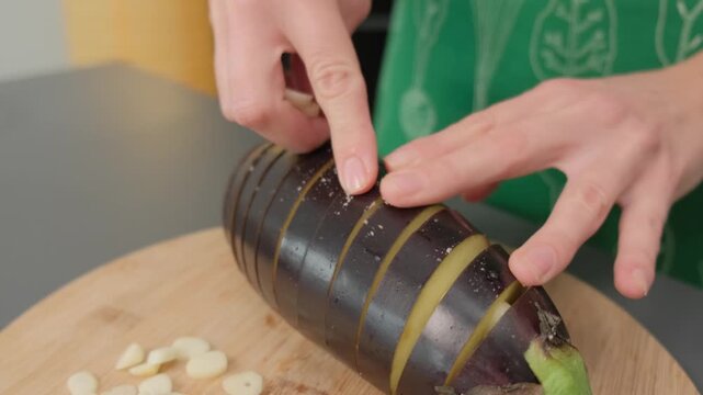 Hands in green clothing slice garlic cloves and arrange them on dark root vegetable rounds on a wooden board during cooking preparation indoors.
