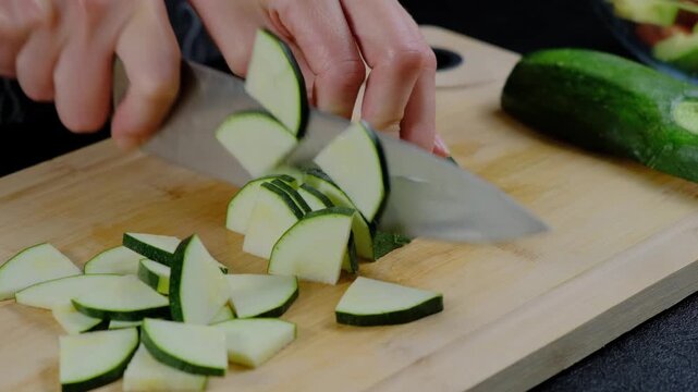 A person slices zucchini on a cutting board with a serrated knife in a Cooking kitchen during daytime.