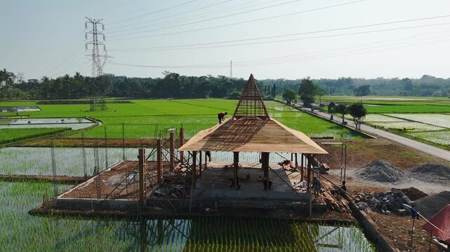 Aerial view of a traditional Javanese joglo structure under construction amidst lush green rice fields with a worker on the roof in Kecamatan Tingkir, Jawa Tengah, Indonesia.