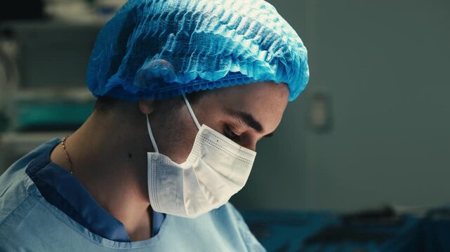 Female surgeon wearing a medical mask, sterile clothing and disposable cap looks down with concentration at the surgical field during a medical procedure.