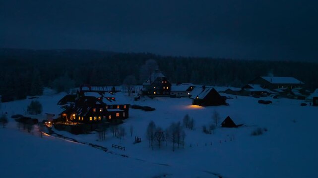 Aerial view of Jizerka village illuminated at dusk with snow-covered landscape and dark winter sky in Korenov, Liberec Region, Czechia.