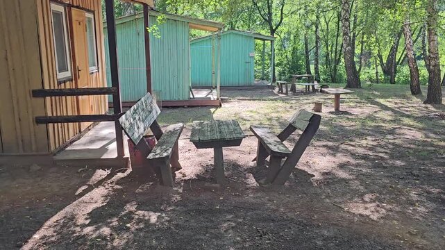 Small wooden cabins with picnic tables in a peaceful forest recreation area surrounded by tall trees on a sunny summer day