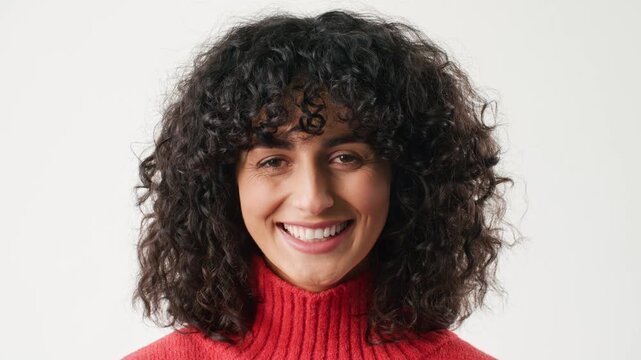 Portrait of cheerful young Caucasian woman with curly dark hair smiling brightly at camera. Happy female wearing red sweater. Natural beauty expression in soft daylight with clean background.