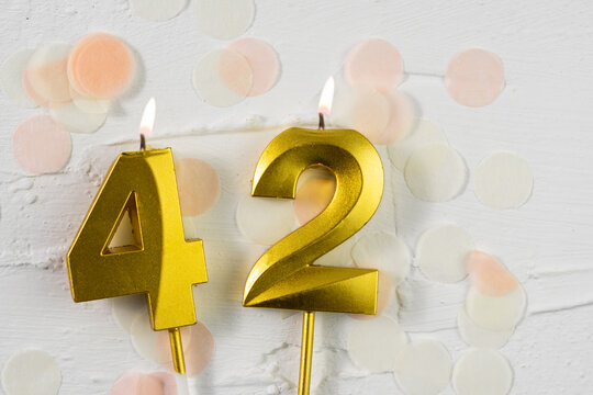 Forty-second anniversary celebration. A golden burning candle in the shape of the number forty-two on a festive white background with confetti and glitter. Festive banner.