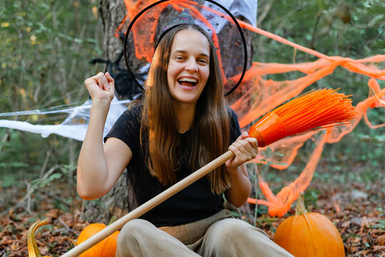 A young woman enjoys Halloween in the woods surrounded by pumpkins. She holds a broomstick and smiles amid decorations like spider webs and orange colors