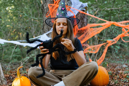 A girl sits cross-legged in the woods wearing a witch hat. She holds a toy spider while pumpkins and Halloween decorations are nearby