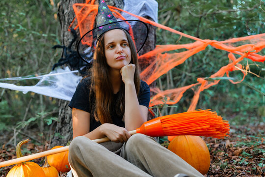 A young woman sits on the ground among pumpkins in a forest. She wears a witch hat and holds a broom. Halloween decorations are in the background