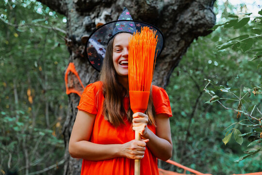 A woman stands in a forest wearing a witch hat and a bright orange dress. She holds an orange broom and smiles while leaning against a tree during a Halloween event