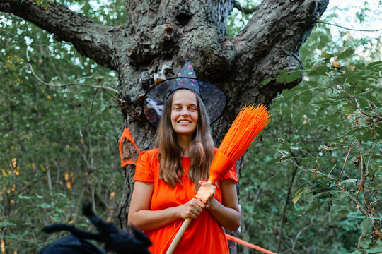 A woman dressed in an orange outfit stands in front of a large tree in a forest. She holds a broomstick and smiles while wearing a witch hat