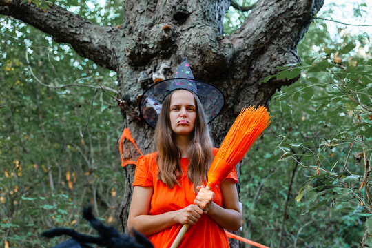 A woman wears an orange dress and holds a broom while standing next to a tree in a forest. Surrounding trees and plants create a Halloween setting