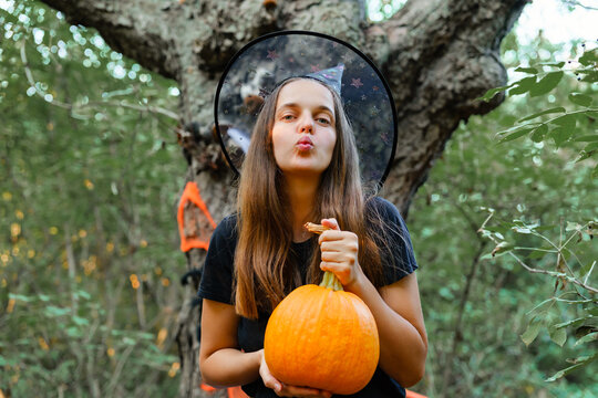 A girl stands outside near a tree, holding a pumpkin and blowing a kiss. She wears a hat and is surrounded by green leaves on an autumn day