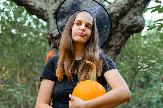 A woman stands in a forest holding a pumpkin. She wears a witch hat and a black outfit. The scene shows autumn leaves and trees in the background