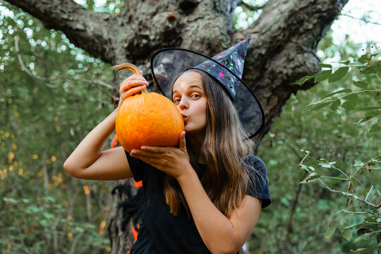 A girl in a witch hat holds an orange pumpkin close and kisses it in a forest. Trees surround her as she celebrates autumn