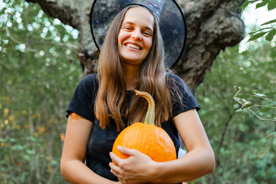 A woman stands in the woods smiling while holding a bright orange pumpkin. She wears a witch hat and enjoys the fall weather surrounded by trees