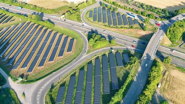 Aerial view of rows of solar panels installed in a highway cloverleaf interchange surrounded by green fields and trees in Tubingen, Baden-Wurttemberg, Germany.