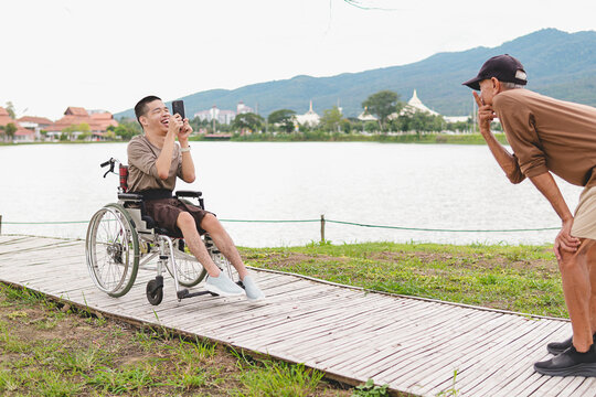 Happy disabled Asian young man on wheelchair using smartphone to take photo of father, joyful family travel, inclusive outdoor lifestyle, caregiving bond, accessibility and handicap empowerment