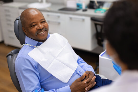 Smiling senior black male patient in dental chair listening to female dentist during friendly consultation at modern dental clinic, reflecting comfort, trust and a positive dental care experience