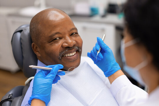 Closeup portrait of smiling senior black male patient holding dental mirror tool, posing with female dentist during checkup appointment