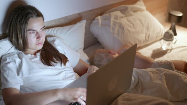 Young Caucasian woman in white shirt using laptop in bed while toddler sleeps beside her, soft lighting creates a cozy atmosphere in the bedroom