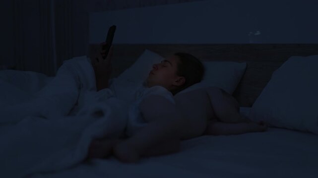 Young woman lying in bed at night, using smartphone while wrapped in white blanket, soft lighting creating a calm atmosphere in a cozy bedroom setting