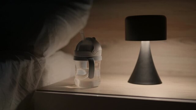 Water bottle with straw and black lamp on bedside table beside white bedding, illuminated softly in a cozy bedroom setting during nighttime