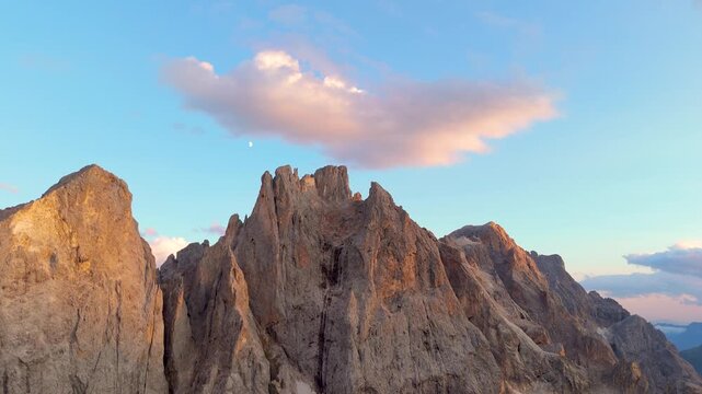 Aerial view of Dolomites mountain peaks illuminated by golden hour light with a pink cloud in the sky in San Martino di Castrozza, Trentino-Alto Adige, Italy.