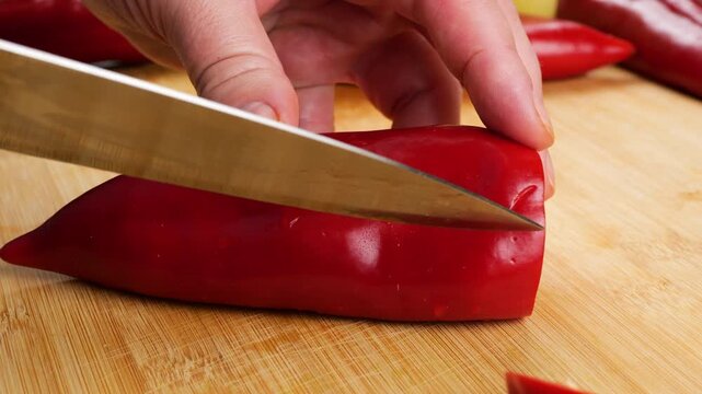 A chef prepares a fresh sweet red pepper on a wooden cutting board, cutting it lengthwise with a sharp knife to remove the white seeds and ribs for a healthy vegetarian dish.