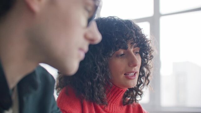 Young curly haired Caucasian woman listening carefully to colleague during discussion. Female looking thoughtful while working together with teammate in bright modern office or classroom.