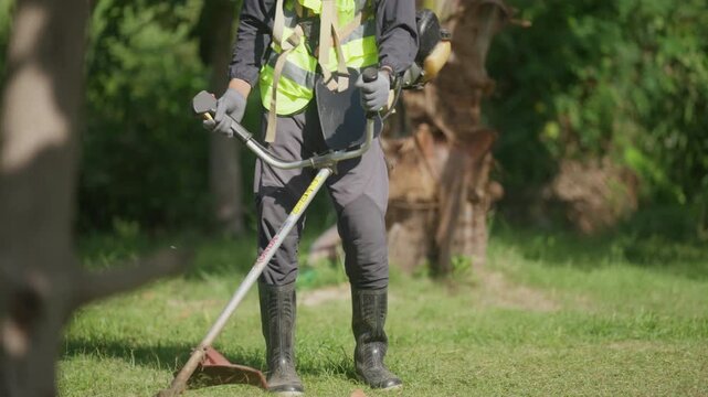 Landscape worker trims overgrown grass with powered equipment in a tropical green space, illustrating routine property maintenance, professional gardening and safe outdoor work.