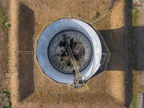 Aerial view of an industrial circular storage tank with a metal walkway and concrete walls surrounded by earthen berms in Chivasso, Piedmont, Italy.