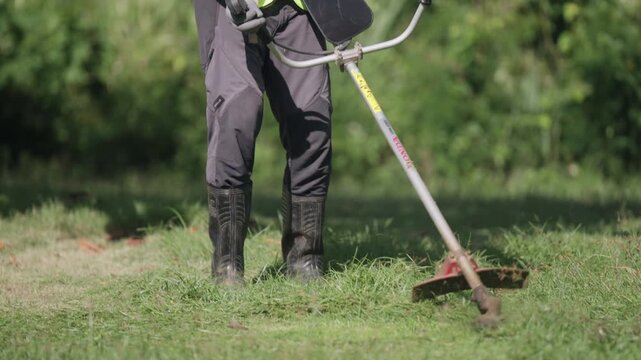 Professional gardener operates a powered brush cutter while maintaining a tropical lawn, wearing protective work gear for safe landscaping, vegetation control and outdoor maintenance.