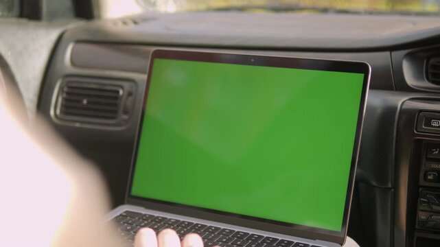 Person sits in a car, using a laptop with a green screen. The laptop is open, resting on their lap. Hands type gently on the keyboard, focused on work. The car's interior shows dashboard