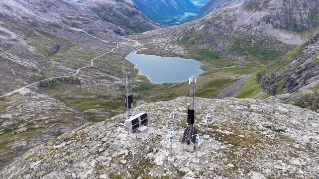 Aerial view of Trollstigen mountain peak with communication towers, solar panels, and wind turbines overlooking a lake and winding road in Andalsnes, More og Romsdal, Norway.