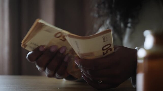 Close-up top view of a Black woman holding and counting a large amount of fifty euro banknotes over a wooden table, representing savings, income, wealth and financial planning.
