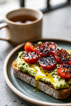 A slice of artisan bread topped with mashed avocado, ripe cherry tomatoes, a sprinkle of black sesame seeds and olive oil.