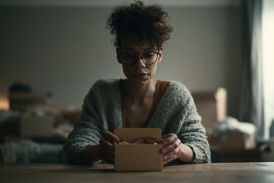 A person sitting at a table, holding a cardboard box containing a personal items. The background is packed with boxes, suggesting a move.