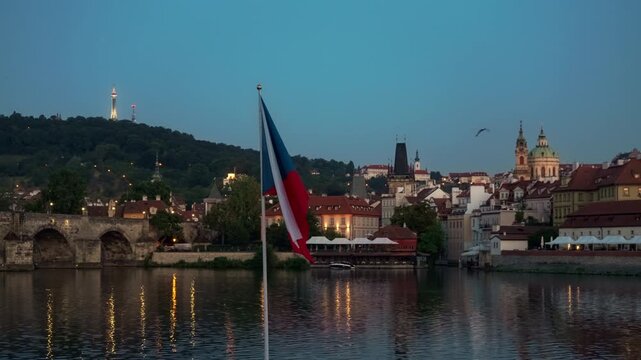 Czech national flag flutters in foreground against Prague skyline at dusk. Illuminated Charles Bridge and Vltava River reflect evening lights. Iconic Czechia architecture represents travel.