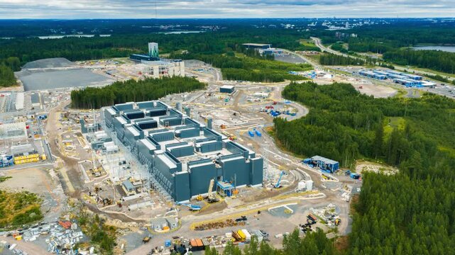 Aerial view of data center construction site surrounded by dense pine forests and lakes under a cloudy sky in Kirkkonummi, Uusimaa, Finland.