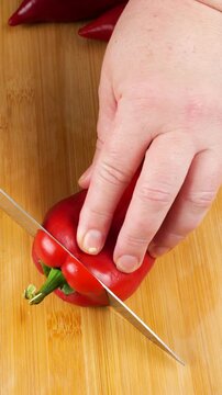 A chef prepares a fresh sweet red pepper on a wooden cutting board, cutting it lengthwise with a sharp knife to remove the white seeds and ribs for a vegetarian dish. Top view. Vertical video.