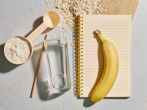 A simple overhead shot of a banana, scoop of protein powder, glass of water, and notebook on a textured surface, suggesting a healthy meal preparation