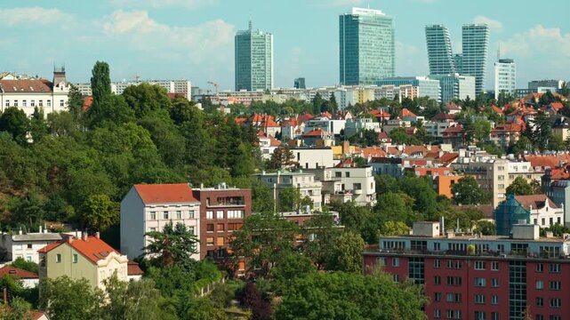 Historic residential houses lie surrounded by green trees in Prague. Modern glass skyscrapers rise in distant business district. Scenic urban view shows city contrast and real estate growth.