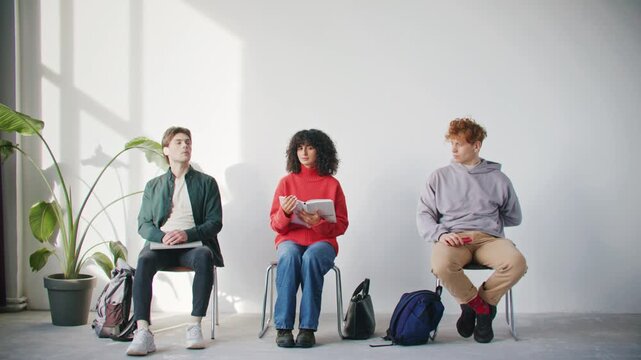 Three young people sitting in waiting room with different interests. Woman reading notebook while two men holding smartphones and looking around. Modern lifestyle. Bright minimalist interior.