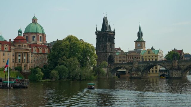 Small tourist boat sails along Vltava River in historic Prague. Old Town Bridge Tower and Charles Bridge span across water. Iconic Czech architecture highlights European travel heritage.