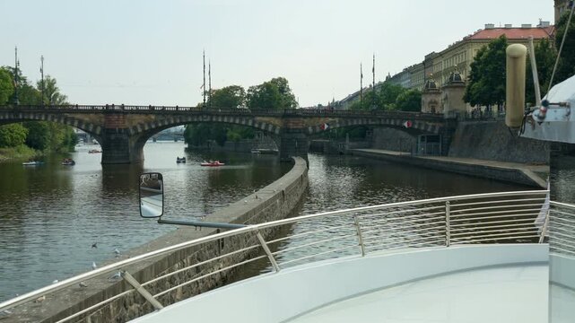 A tourist boat approaches Smichov Lock entrance on Vltava River in Prague, Czechia. Historic stone bridge spans across water. Czech waterway engineering shows European river cruise travel scene.