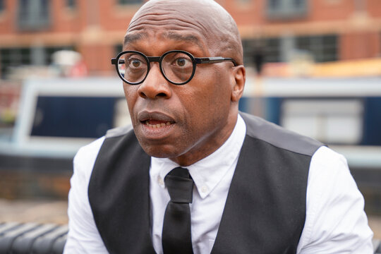 Man in glasses looks surprised while sitting outdoors by a canal in a city during daytime