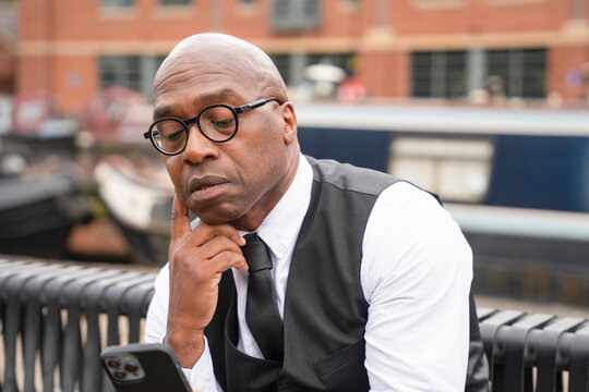 Man in a suit checks his phone while sitting on a bench near a canal in an urban area during the day