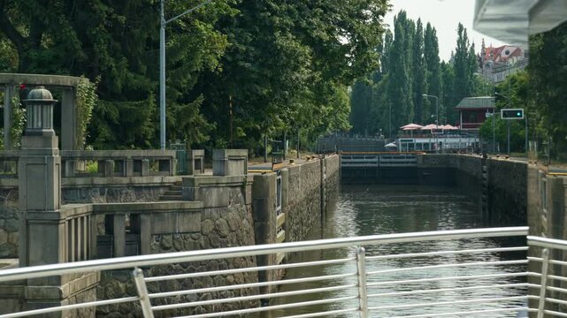 A tourist boat enters Smichov Lock chamber on Vltava River in Prague, Czechia. Stone walls guide river navigation toward closed gates. European waterway hydraulic engineering travel scene.